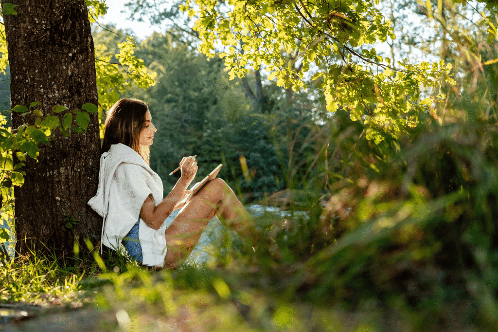 A woman leans against a tree and journals.