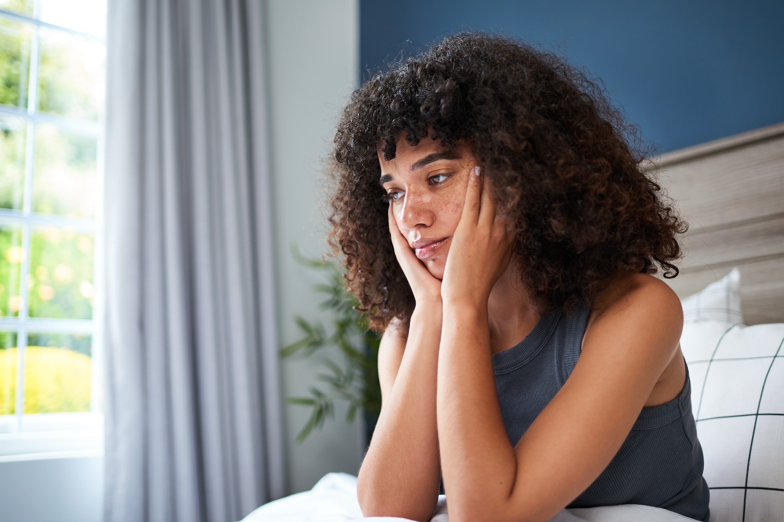 Young Woman Sitting Thoughtfully on a Bedroom Bed With Natural Light
