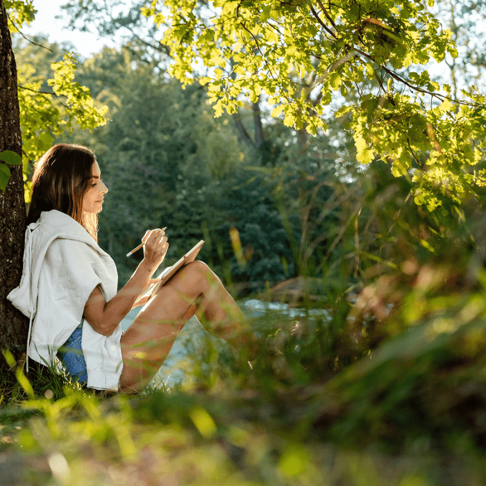 A woman leans against a tree and journals.